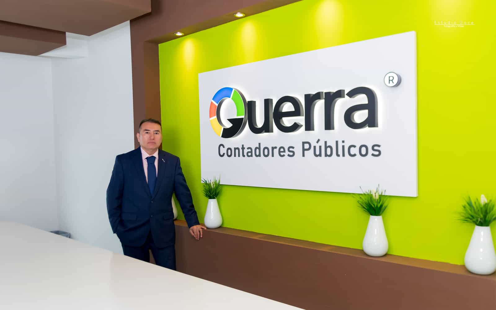 El C.P.C. Dr. Jesús Guerra posando en la recepción de la oficina junto al logotipo de Guerra Contadores Públicos